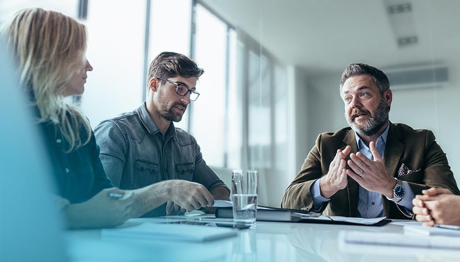 Zwei Männer und eine Frau sitzen in einem Besprechungsraum und gestikulieren bzw. diskutieren offensichtlich über Dokumente, die vor ihnen ausgebreitet sind.