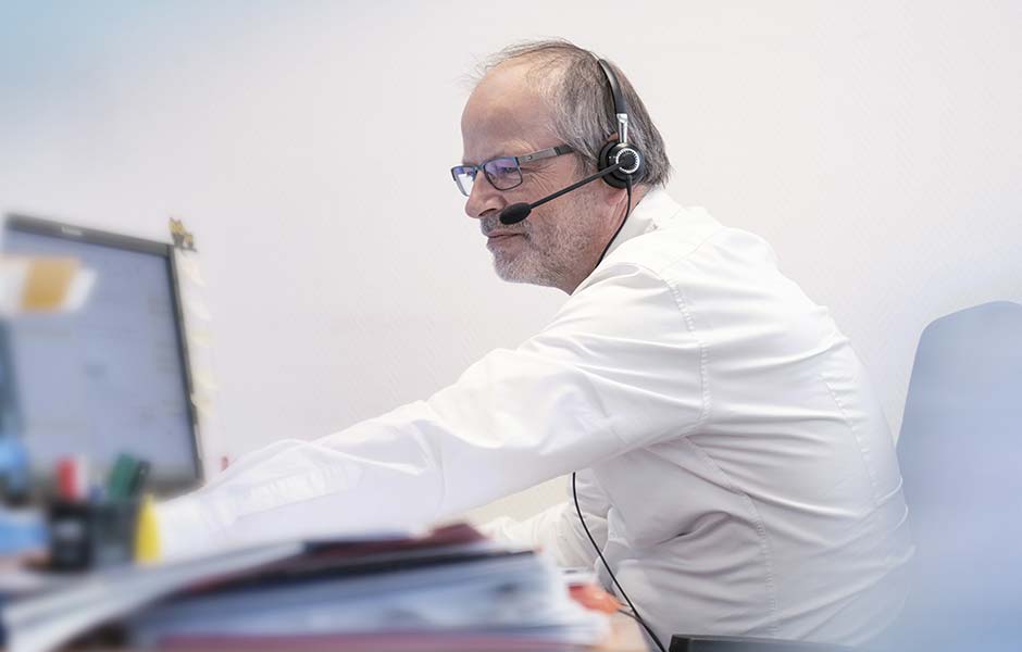 Ein Mann mit Headset sittz in einem Büro. Man sieht einen Aktenstapel, einen Bildschirm und die Rückenlehne seines Stuhls.